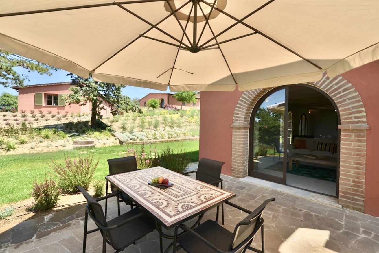Eine Terrasse im Freien mit Tisch und Stühlen, einem großen Sonnenschirm und Blick auf einen Garten sowie Gebäude im Hintergrund.