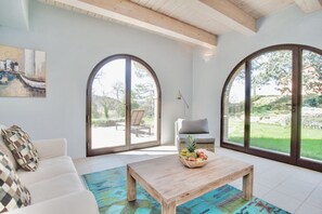 Un salon spacieux avec un canapé blanc, une table basse en bois sur laquelle sont disposés des fruits, et de grandes fenêtres en arc de cercle offrant une vue sur l’extérieur.