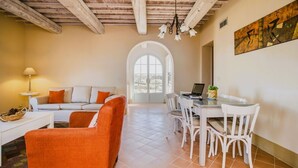 Un vaste espace de vie comprenant un canapé blanc, un fauteuil orange et une table à manger avec des chaises. Un ordinateur portable est posé sur la table, et une fenêtre offre une vue sur les arbres.