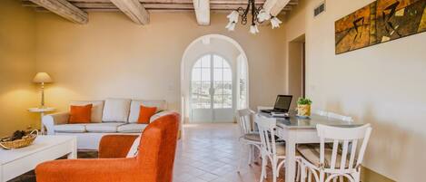 A spacious living area with a white sofa, orange armchair, and dining table with chairs. A laptop is on the table, and there is a window with a view of trees.