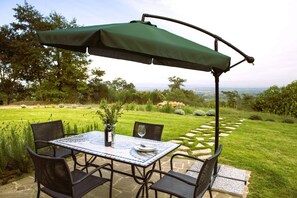A patio with a table and chairs, a green umbrella, and a view of a landscaped garden. - Elegant ground floor apartment of 92 m2, composed of a large living room with fireplace, kitchen complete with dishwasher, microwave oven, electric oven, coffee machine, kettle, fridge and freezer, internal and external dining area, a double bedroom with (Trequanda)