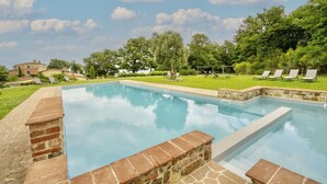 Une piscine rectangulaire bordée de pierres, entourée d’une pelouse agrémentée d’arbres et de quelques chaises longues.