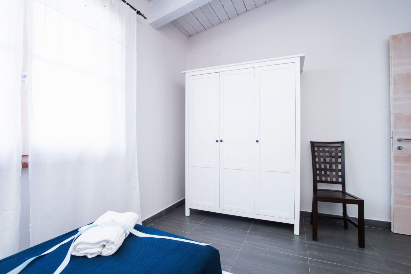 A room with a bed, a white wardrobe, a wooden chair, and a window with sheer curtains.