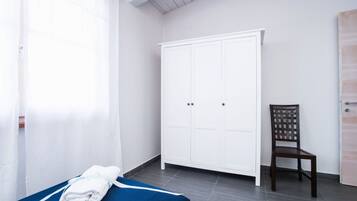 A room with a bed, a white wardrobe, a wooden chair, and a window with sheer curtains.