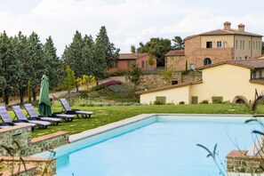 A swimming pool with lounge chairs and a green umbrella, surrounded by a garden and a stone building.