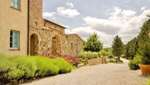 Un bâtiment en pierre avec une allée de gravier et un jardin agrémenté d’arbustes verts et de fleurs roses.