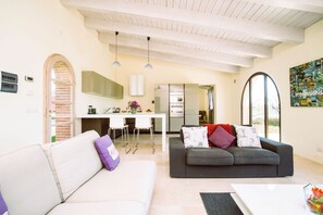 Ein modernes Wohnzimmer mit einem weißen Sofa, einem grauen Sofa und einem Couchtisch. Im Hintergrund ist eine Küchenzeile mit einem Esstisch und Stühlen zu sehen.