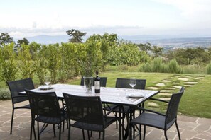 Eine Terrasse im Freien mit einem für vier Personen gedeckten Tisch, umgeben von Stühlen, mit Blick auf einen angelegten Garten und ferne Hügel.