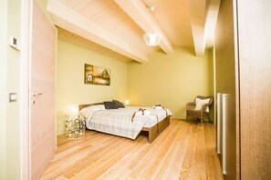 Un dormitorio con techo de madera, una cama con colcha blanca y marrón a cuadros, una silla de mimbre y una mesita de noche con lámpara.