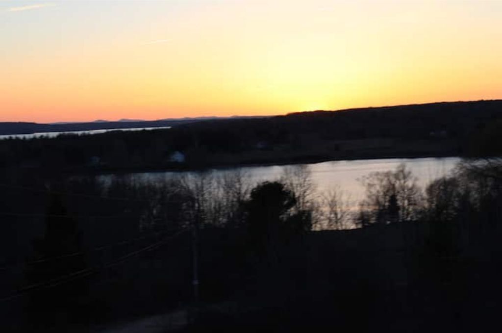 A sunset over a body of water with silhouettes of trees and a distant horizon.