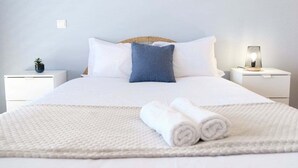 A neatly made bed with white bedding, a blue pillow, and rolled white towels on a textured white rug. - GuestReady - Charming Caniço Retreat (Santa Cruz)