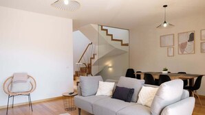 A modern living room with a grey sofa, a wicker chair, a dining area with a wooden table and chairs, and a staircase with wooden steps. - GuestReady - Charming Caniço Retreat (Santa Cruz)