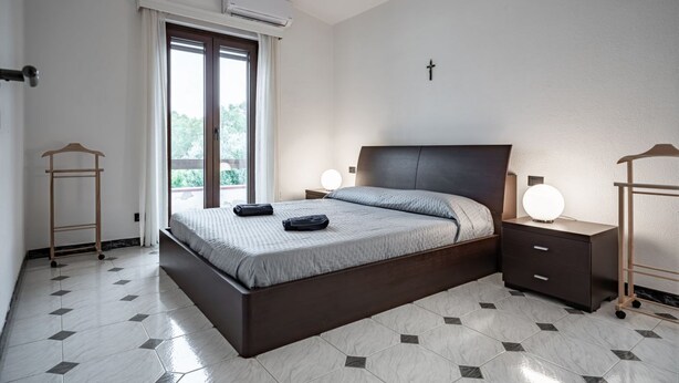 Un dormitorio con cama, mesitas de noche, una cruz en la pared y un ventanal con cortinas.