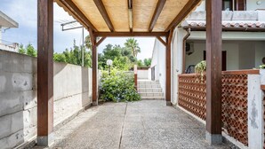 Un espacio al aire libre techado con piso de concreto, una pérgola de madera y vista a un edificio residencial con escaleras.