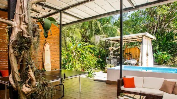 An indoor space with a ping pong table, a sofa, and a pool area visible through large windows.