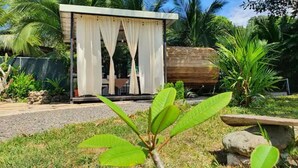 A small, open-air structure with a thatched roof, surrounded by lush greenery and a palm tree.