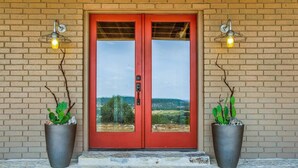 A red double door with glass panels, flanked by two potted plants and two wall-mounted lights. - *New| Stunning Views| Sleeps 10 (Fredericksburg)