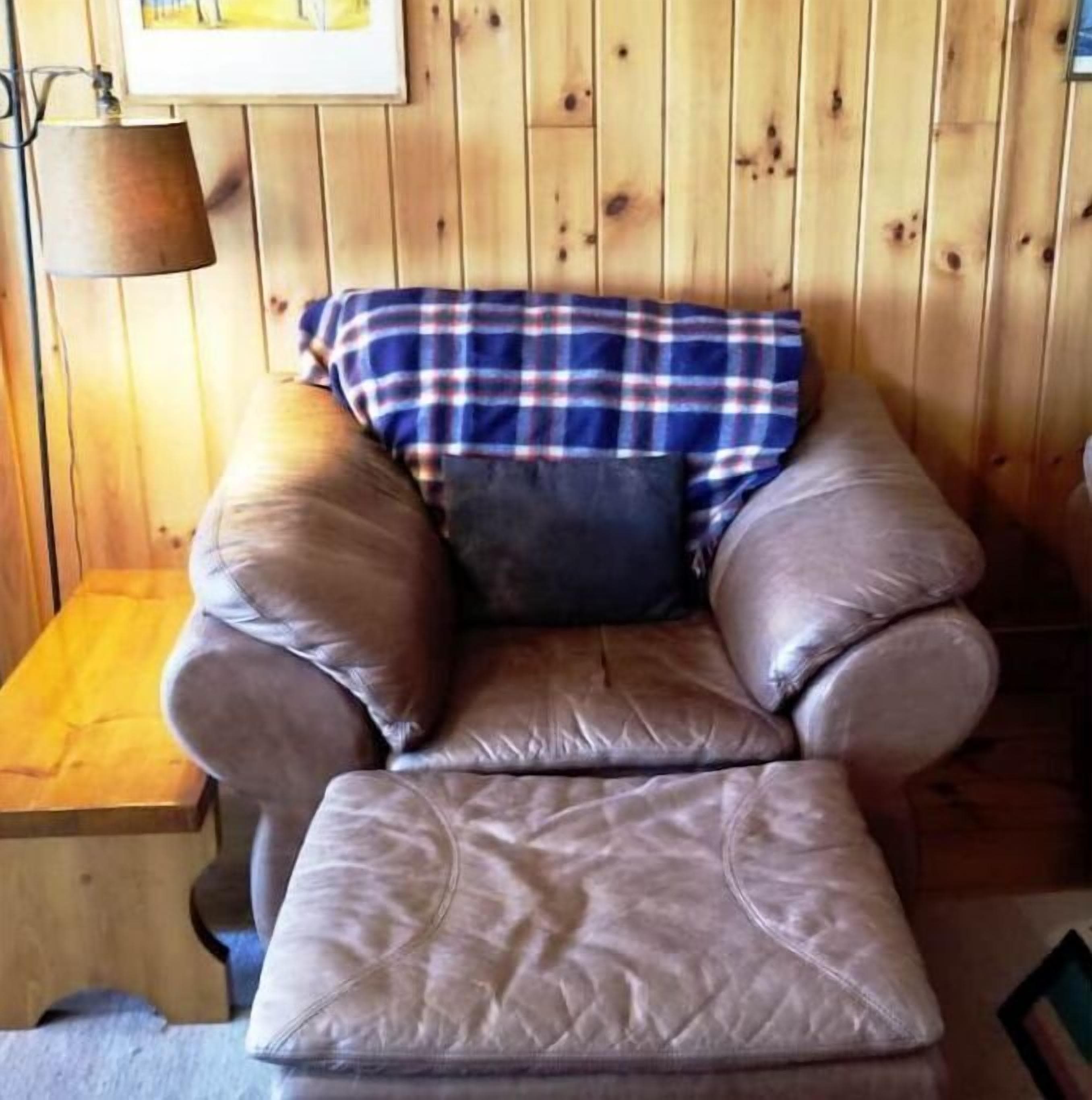 A cozy room with a leather armchair and ottoman, a plaid throw blanket, and a lamp on a wooden wall.