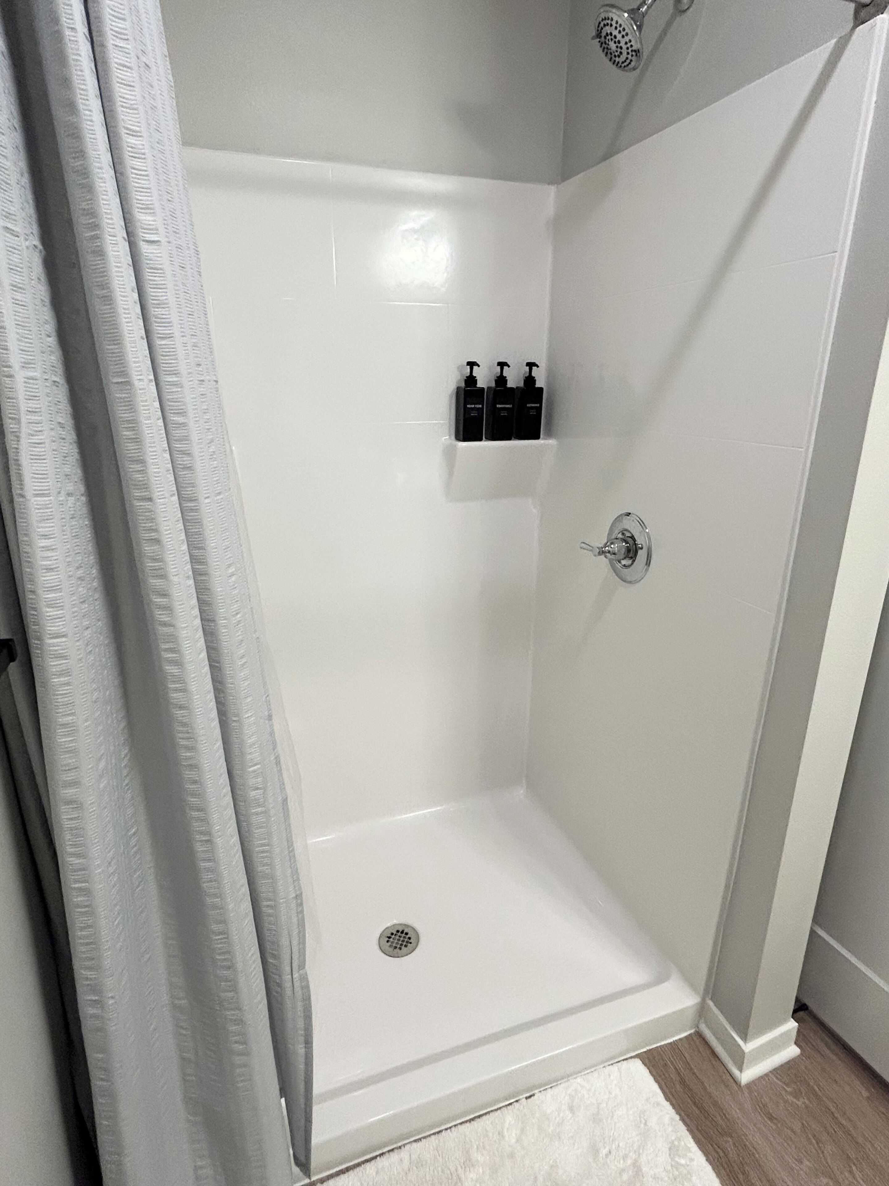 A shower with a white tiled wall, a showerhead, and a drain.