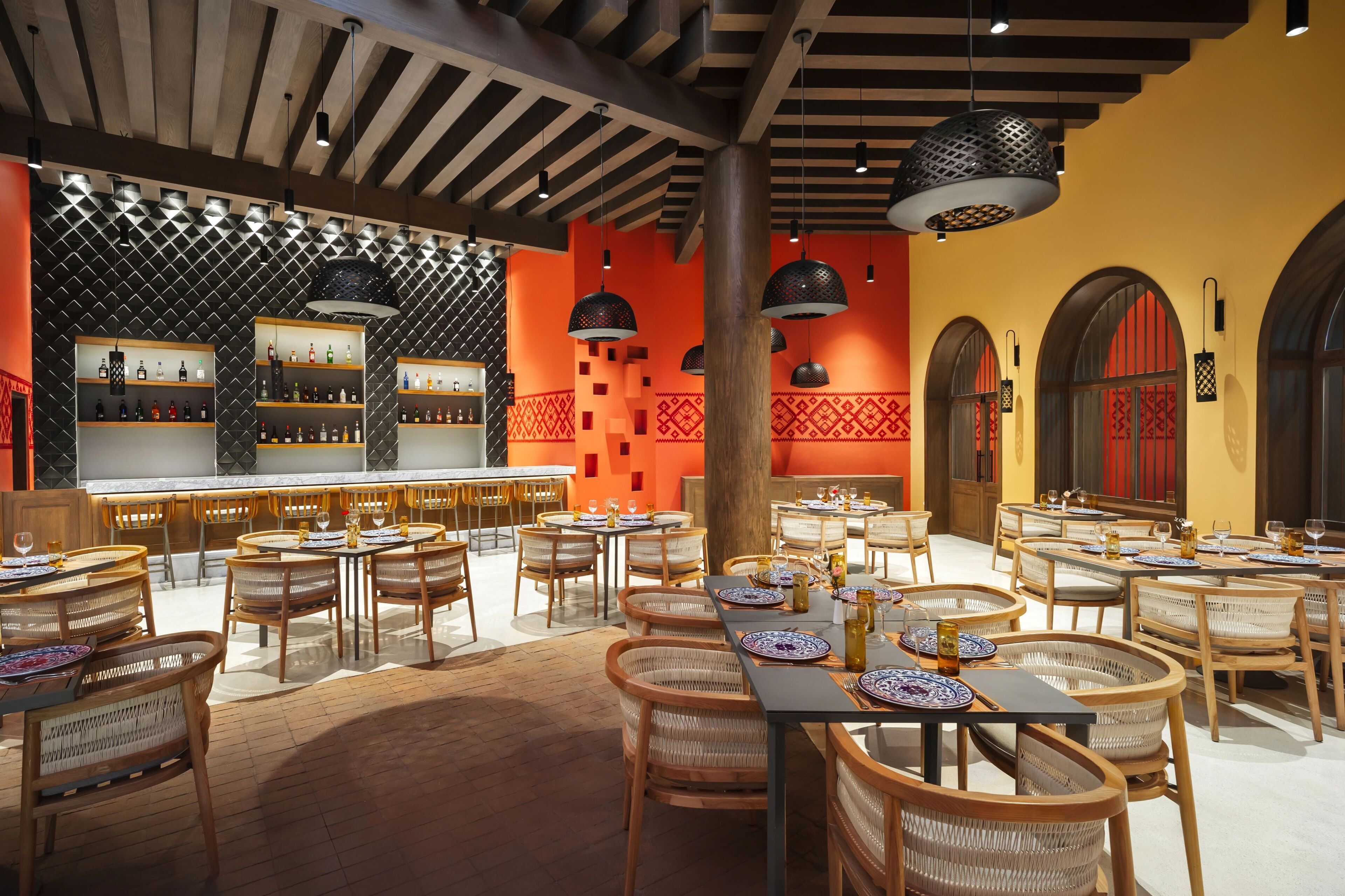 A restaurant with wooden tables and chairs, a bar area with shelves of bottles, and a patterned wall.