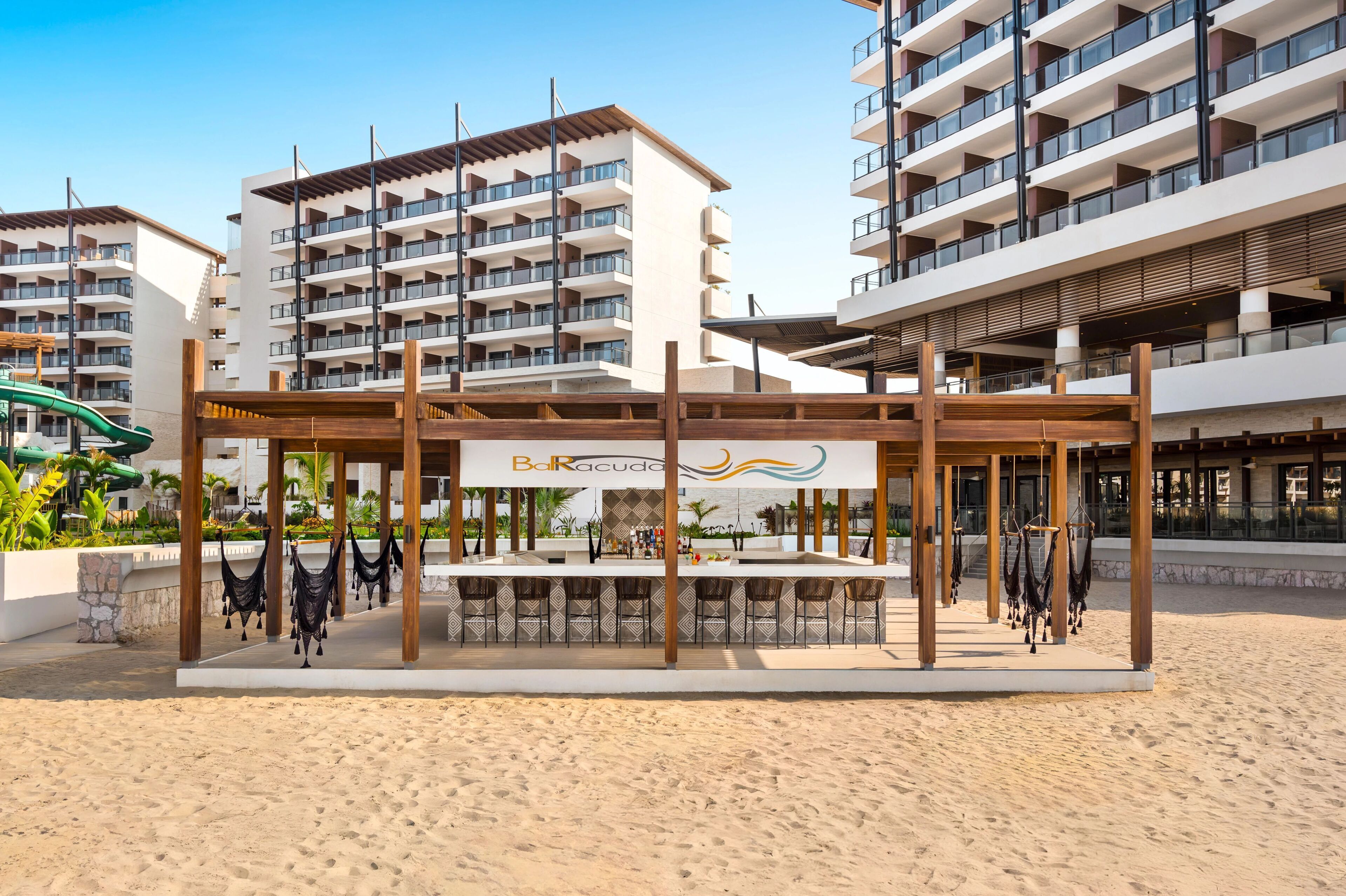 A beachside bar area with a sign reading "Bar Racuda," surrounded by beach chairs and a sandy floor.