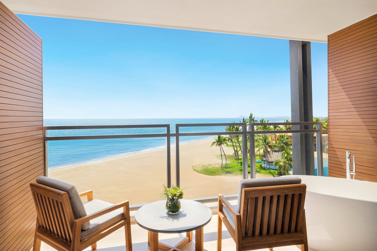 Un balcon donnant sur une plage, deux chaises en bois, une petite table et une plante en pot.