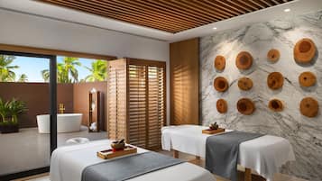 A spa room with two massage tables, a wooden slatted partition, a marble wall with wooden bowls, and a view of palm trees.
