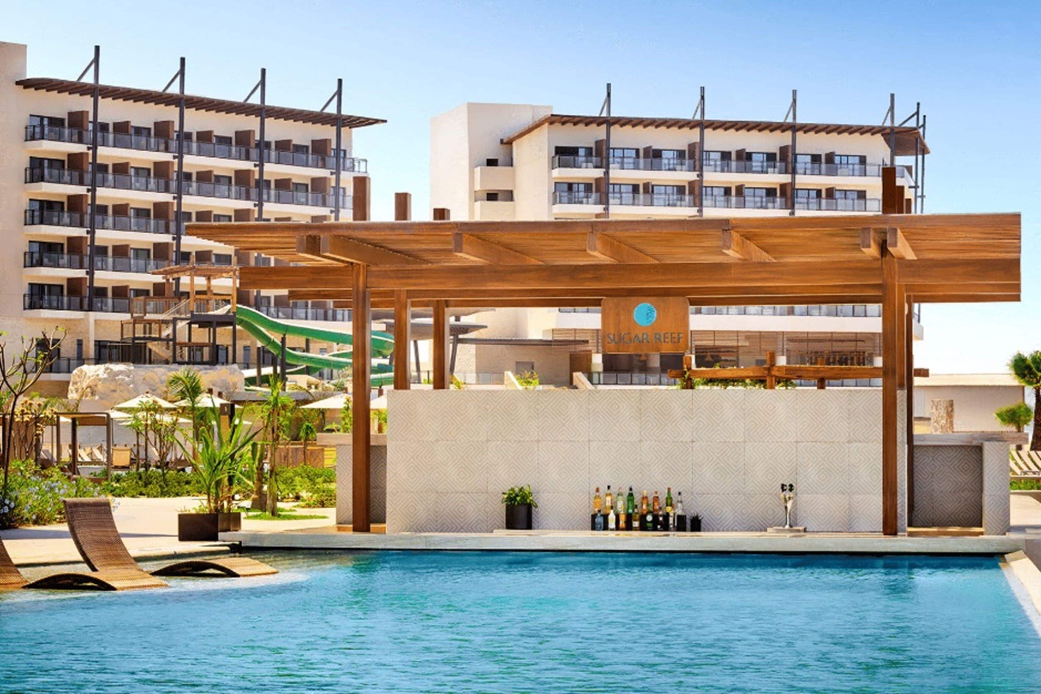 Un área de piscina con una pérgola de madera, un bar con un letrero que dice "Sugar Reef" y un tobogán al fondo.