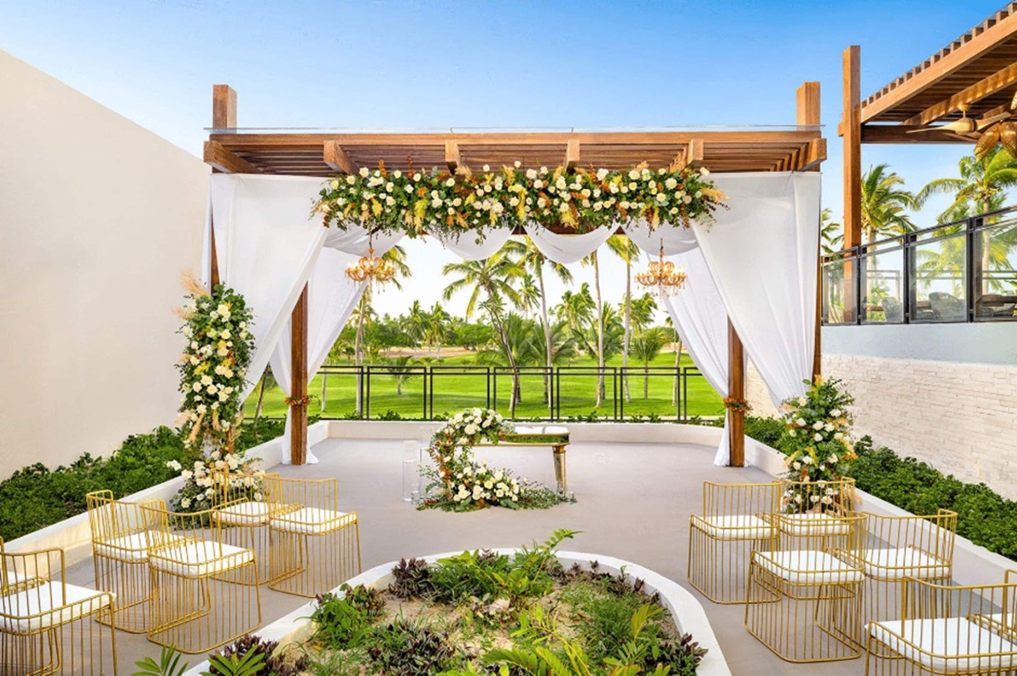 An outdoor wedding setup with white drapes, floral arrangements, and gold chairs.