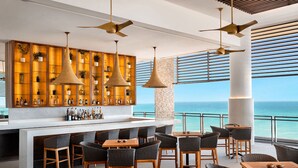 Un bar moderne avec vue sur l’océan, doté de mobilier en osier, d’un éclairage suspendu et d’une grande variété de bouteilles.