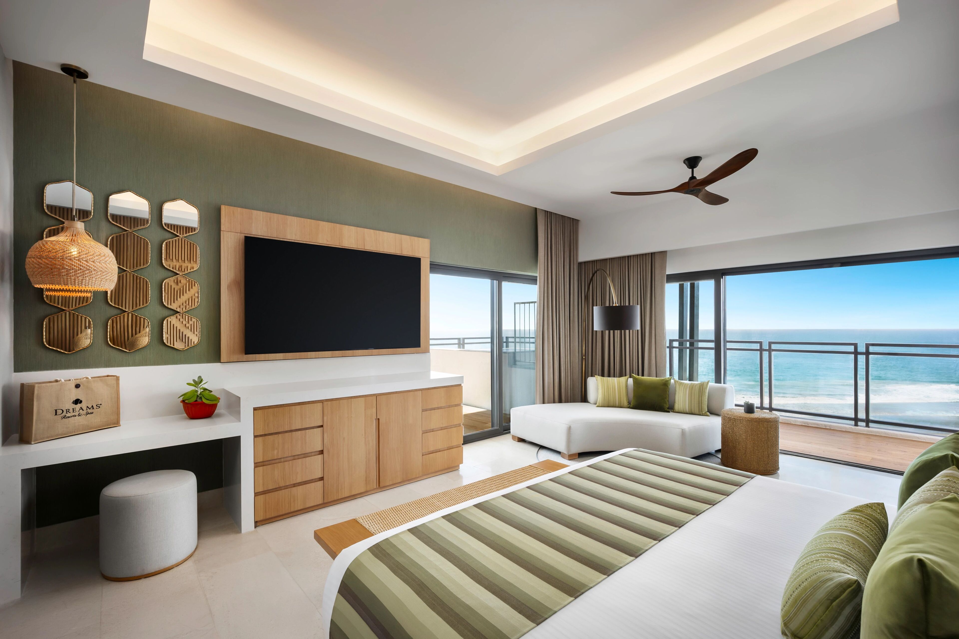 A modern hotel room with a large bed, a TV mounted on the wall, a seating area with a view of the ocean, and a balcony with a railing.