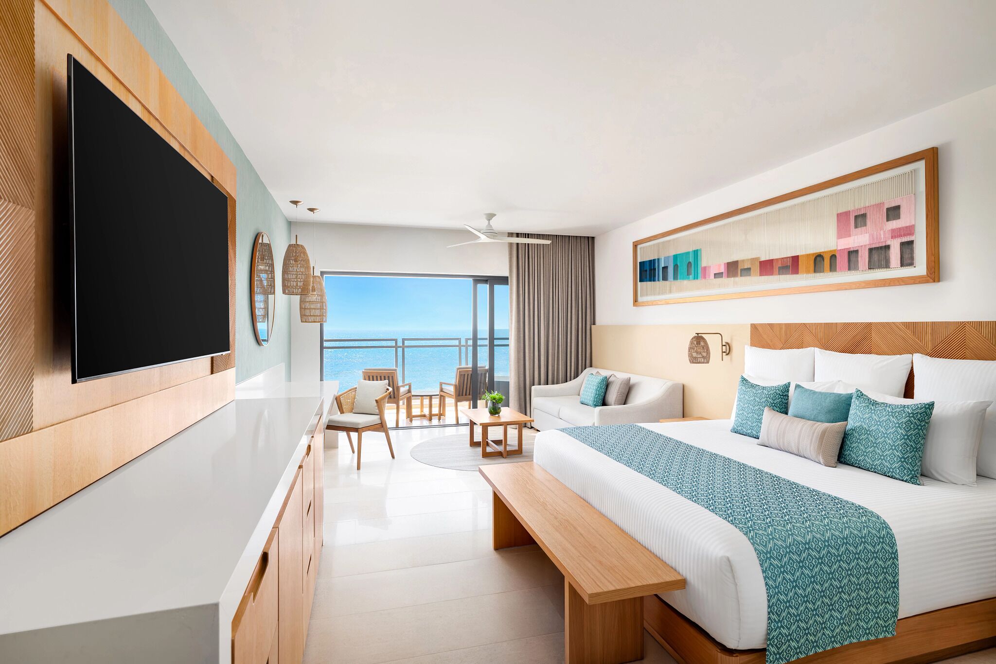 A modern hotel room with a large bed, a flat-screen TV, a balcony with a view of the sea, and a dining area with a table and chairs.