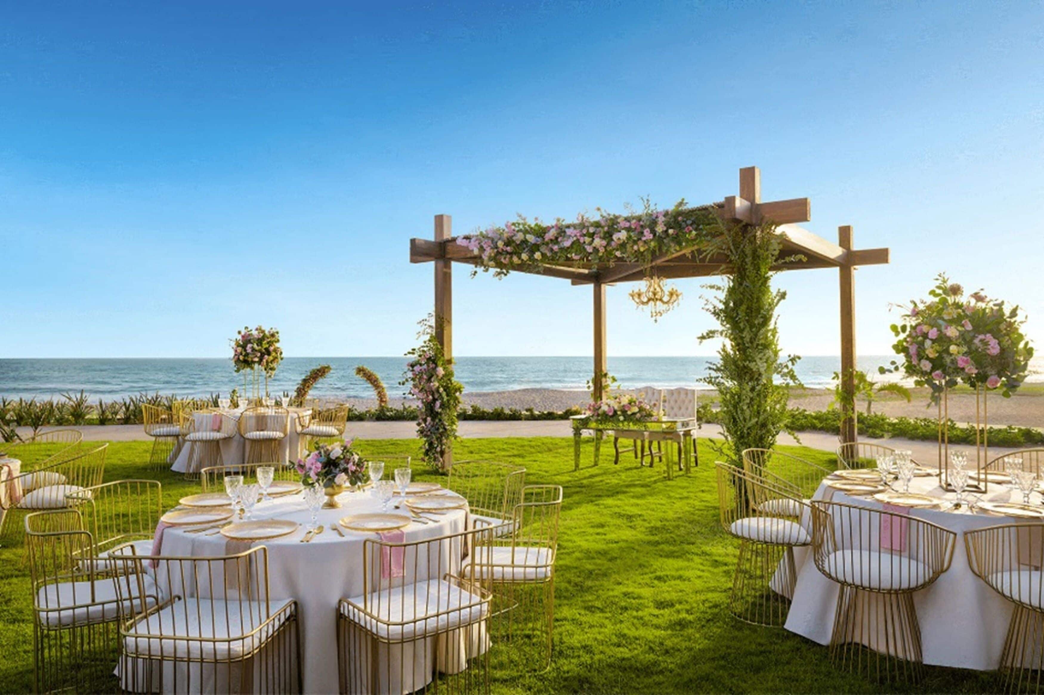 Une cérémonie de mariage en plein air, avec des tables et des chaises disposées sur une pelouse près de la mer, et une arche en bois agrémentée de décorations florales.