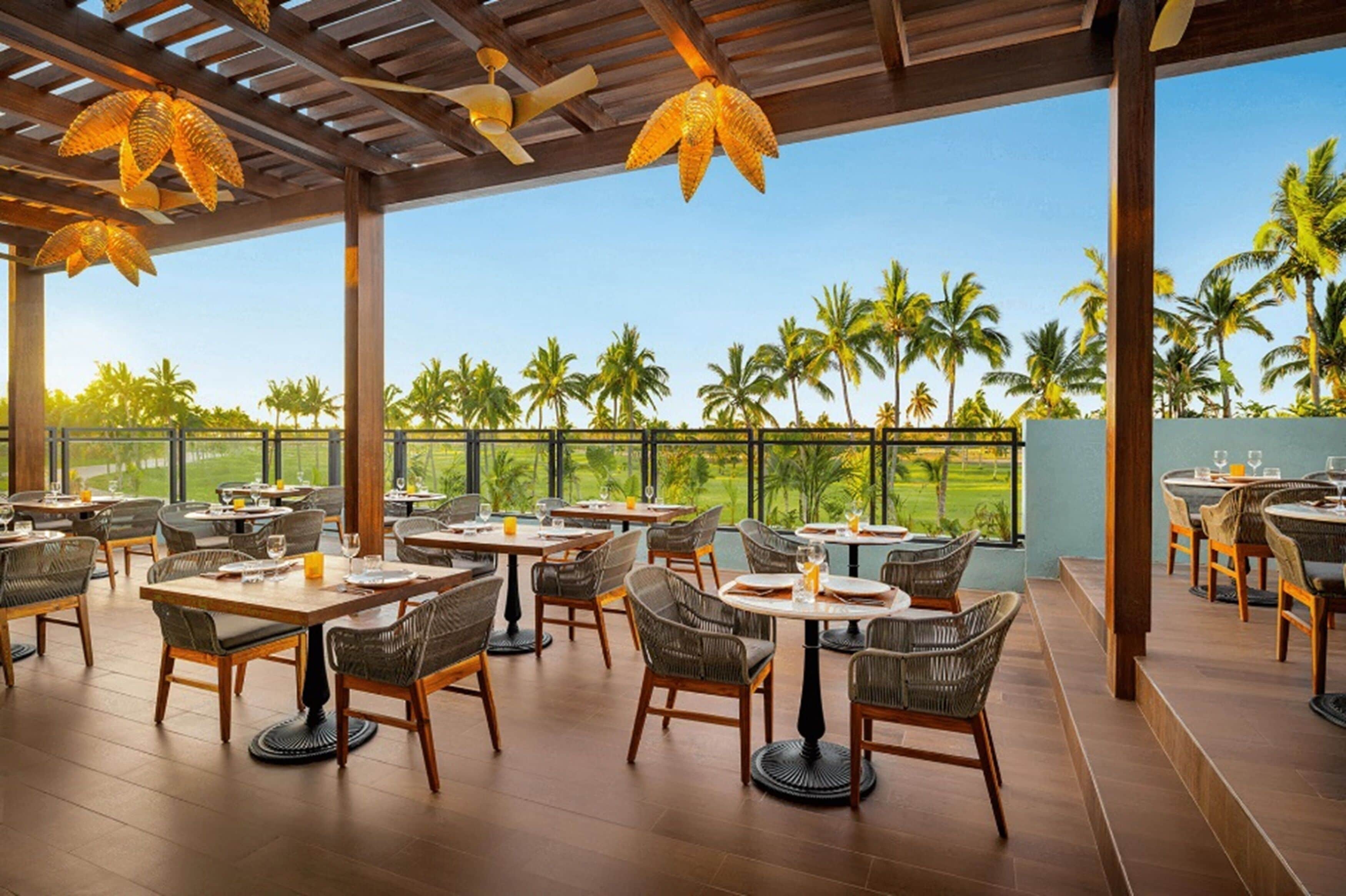An outdoor dining area with wicker chairs and tables, overlooking a lush landscape with palm trees.