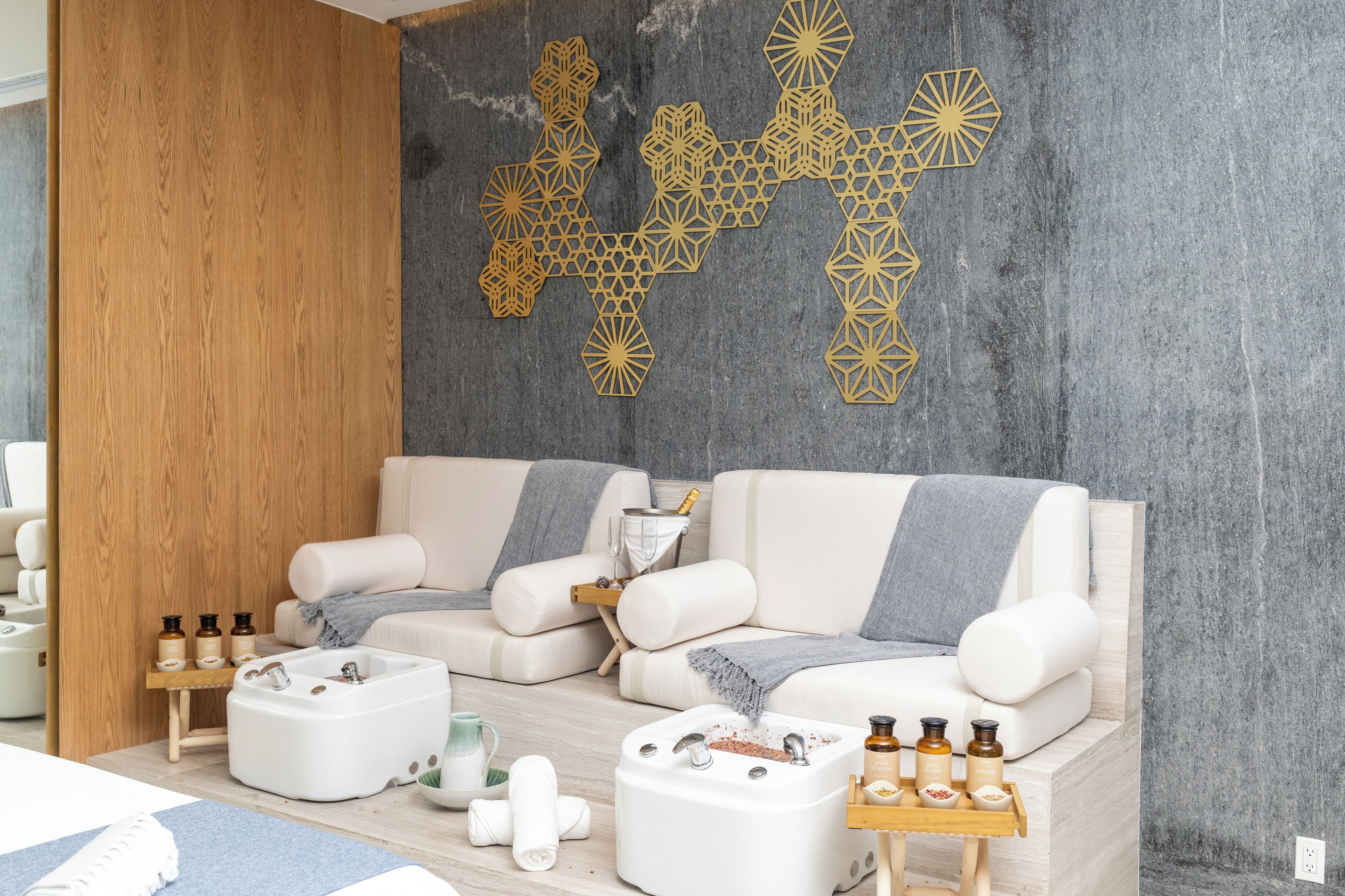 A spa room with two white sofas, a wooden side table with bottles, a small sink, and a decorative wall piece.