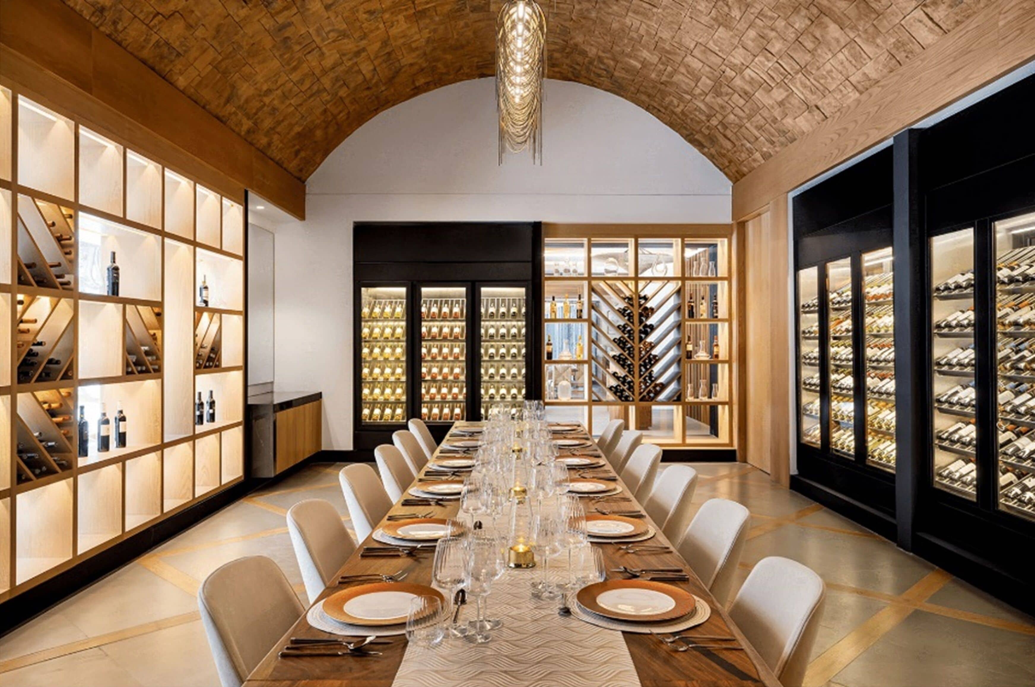 A long dining table set for a formal meal, flanked by wine racks and shelves with bottles.