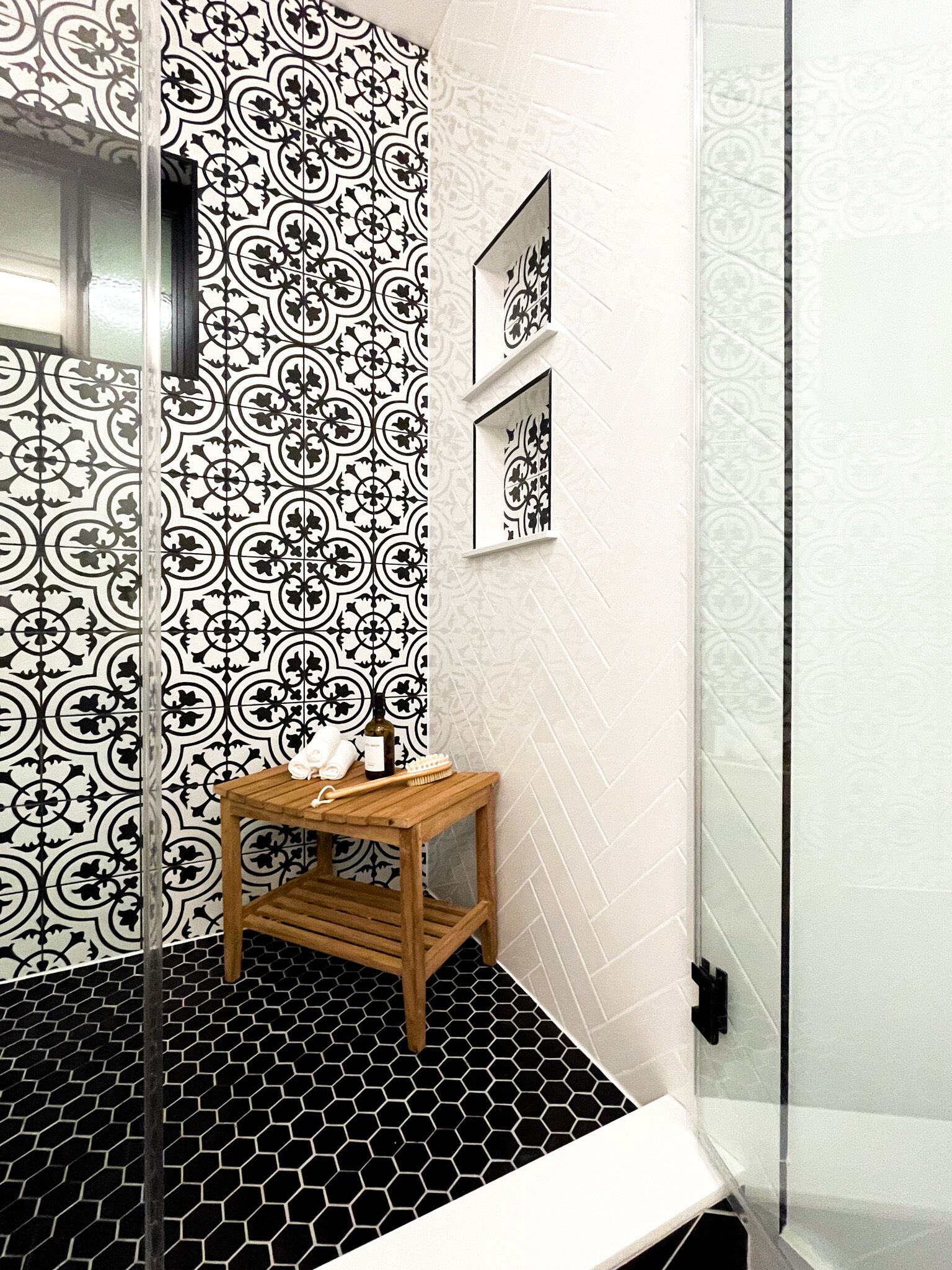 A bathroom with black and white patterned tiles, a wooden vanity, a shower, and a small window.