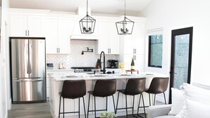 A modern kitchen with white cabinets, a stainless steel refrigerator, a central island with bar stools, and a dining area with a sofa and coffee table. - Modern 2-Bed Family Haven 🏡 Pet-Friendly + EV Charging 🐾⚡ (San Jose)
