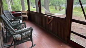A covered porch with wicker chairs and a small table, overlooking a lake and forested area.
