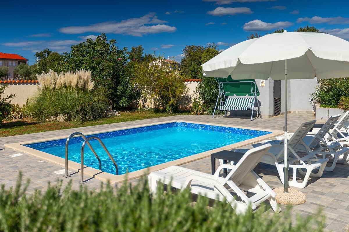 Une piscine rectangulaire à l’eau d’un bleu limpide, entourée d’une terrasse carrelée. On y trouve des chaises longues blanches et un parasol blanc qui offre de l’ombre.