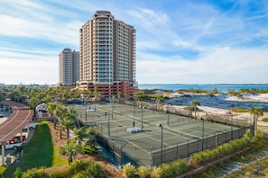 Un court de tennis entouré d’une clôture, avec un espace de stationnement et un bâtiment en arrière-plan.
