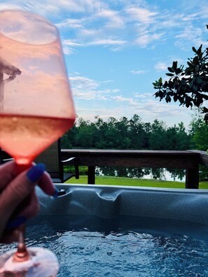 Una mano sosteniendo una copa de vino sobre un jacuzzi con un deck de madera y follaje al fondo.