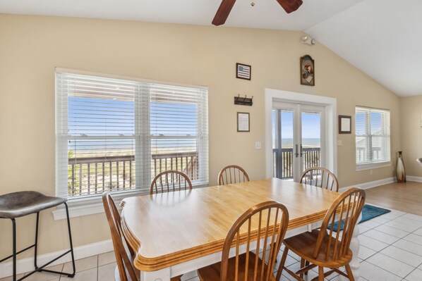 A dining area with a wooden table and chairs, large windows with blinds, a ceiling fan, and a view of the outdoors.
