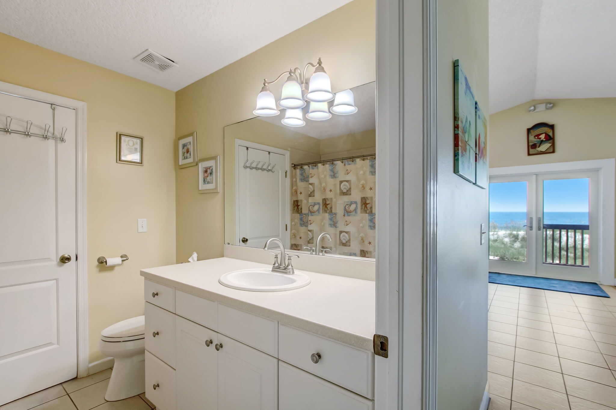 A bathroom with a white sink, mirror, and light fixture. There is a toilet and a door leading to a balcony with a view.
