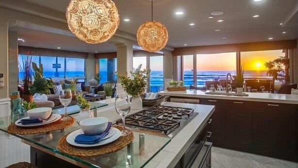 Una cocina moderna con una isla de vidrio, lámparas colgantes con motivos florales y vista al océano al atardecer.