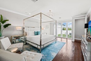 A modern bedroom with a four-poster bed, a sitting area with a chair and a small table, and a large window offering a view of the outdoors.