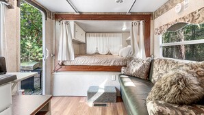 A compact RV interior with a bed, a sofa, a small table, and a window with a view of trees. - River Retreat RV Campground ~ Gym / Game Room / Sauna (Jacksonville)