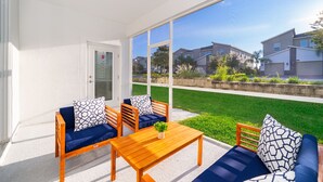 Eine überdachte Terrasse mit einem Holztisch und blau gepolsterten Sitzgelegenheiten, mit Blick auf eine gepflegte Rasenfläche und benachbarte Häuser.