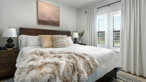 A bedroom with a large bed, two bedside tables, a painting above the bed, and a view through the window.