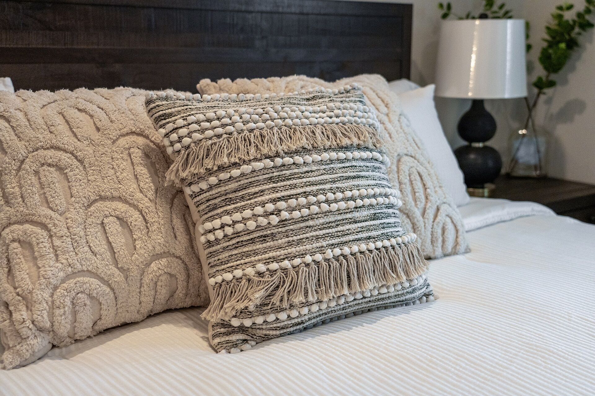 Un lit recouvert d’une couverture beige à texture et agrémenté de deux coussins décoratifs ornés de franges et de détails en perles.